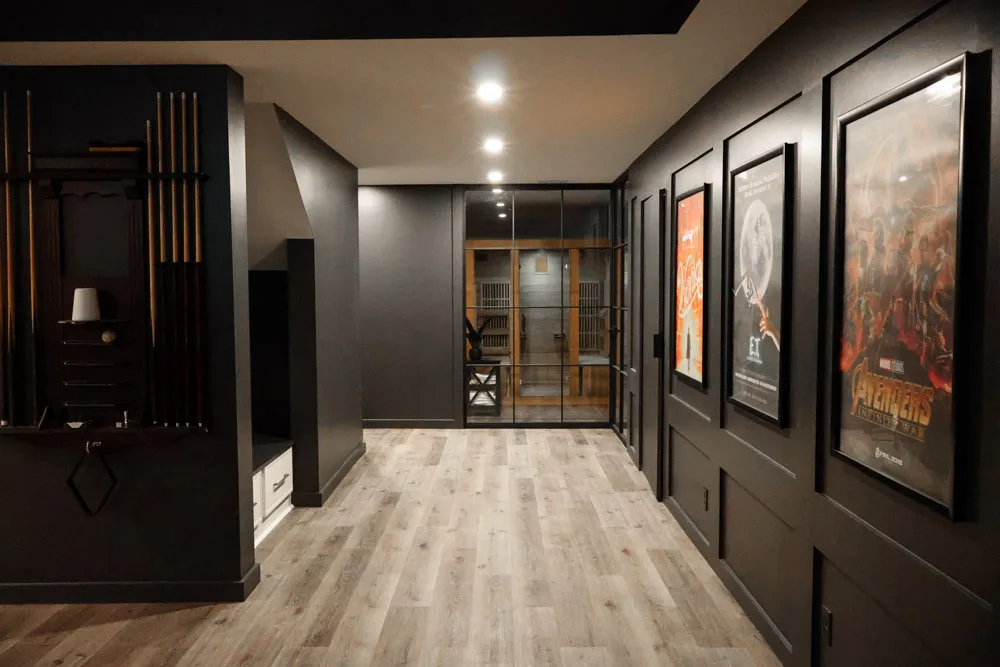 Dim hallway of a home theater with dark walls, wooden floor, framed movie posters on the right, and a glass door at the far end.
