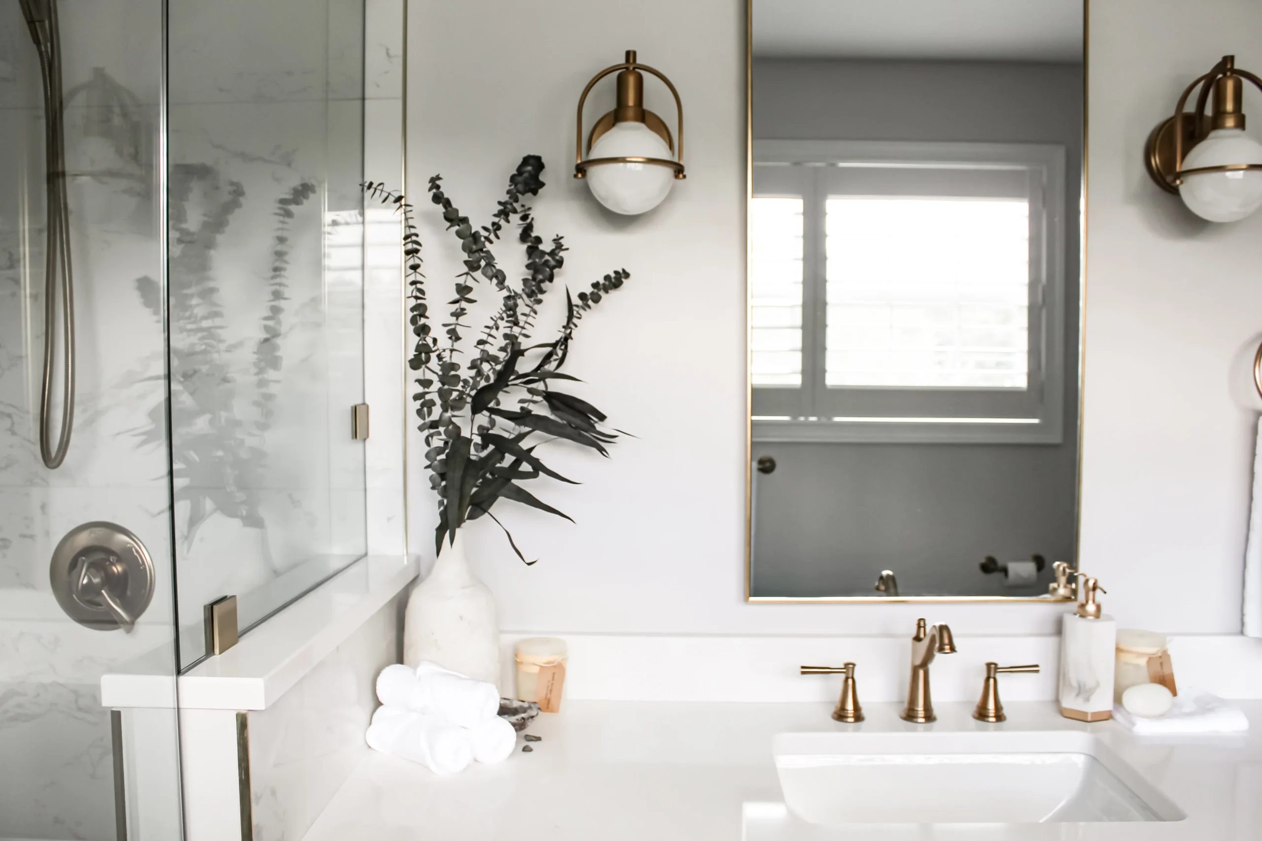 Modern bathroom vanity with white countertop, gold faucets, and a large mirror; a vase with eucalyptus on the counter nearby.