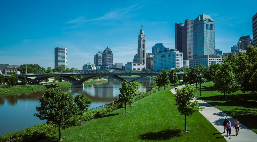 City skyline over a river with a curved bridge, green park, and a sunny blue sky; three walkers on a path in the foreground