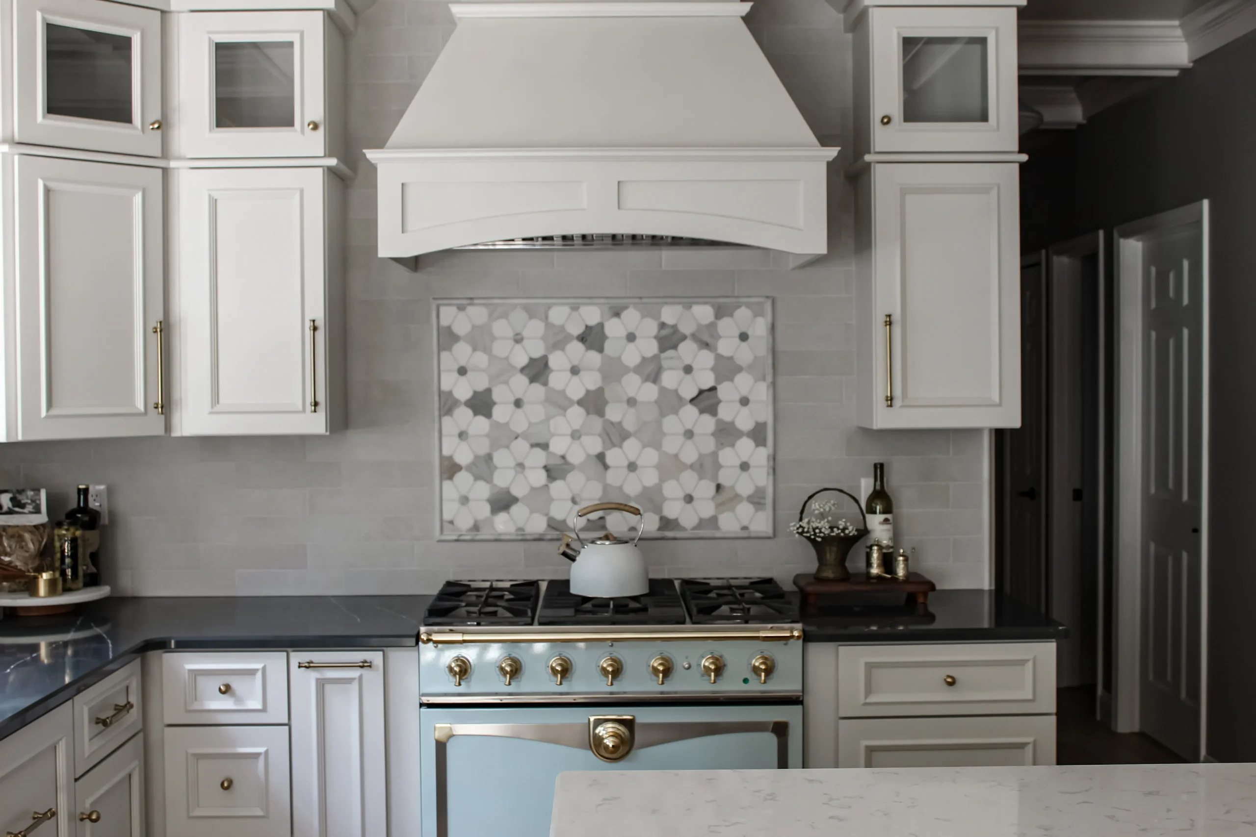 White kitchen with a large range hood above a floral tiled backsplash and brass hardware on cabinets and stove doors.
