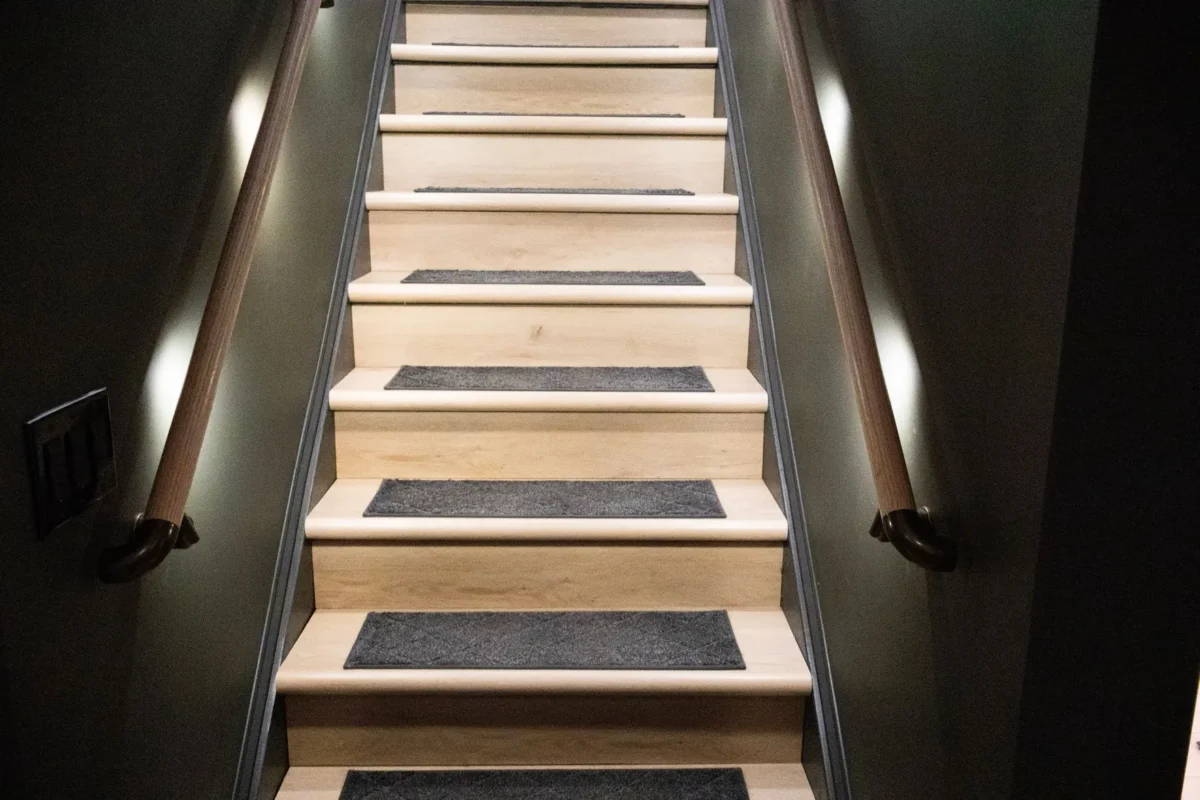 Indoor wooden staircase with dark carpet runners on each step and brown handrails on both sides, viewed from the bottom looking up