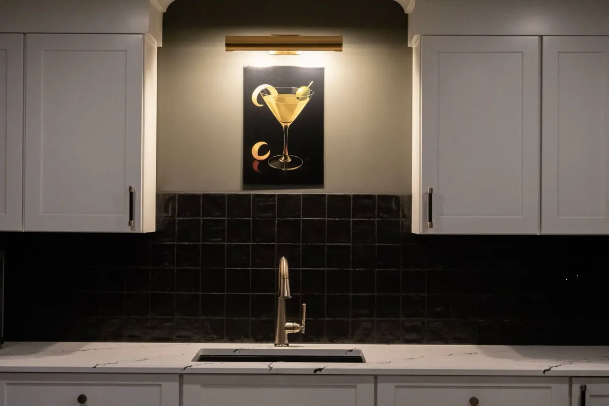 White kitchen cabinets with a glossy black tiled backsplash and a centered wall art depicting a gold cocktail under a warm spotlight above the sink area.