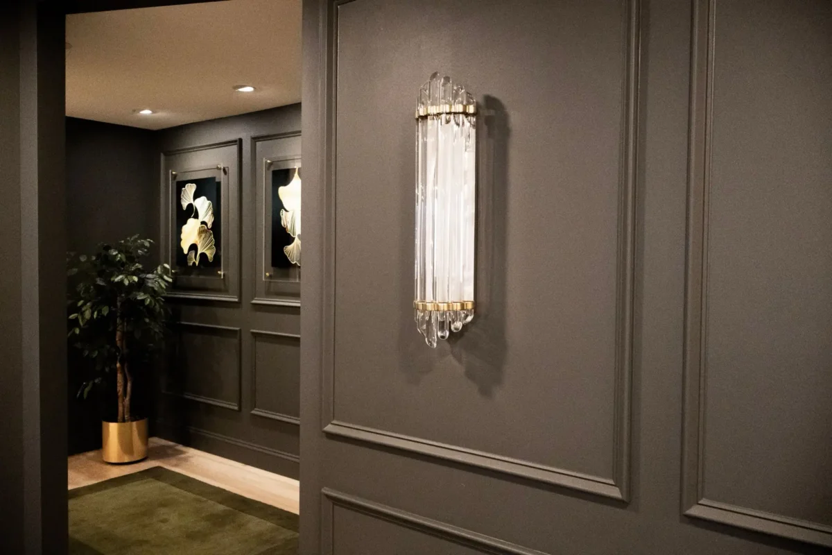 Gray paneled hallway with a crystal wall sconce, framed botanical art, and a gold planter with a green plant in the corner.
