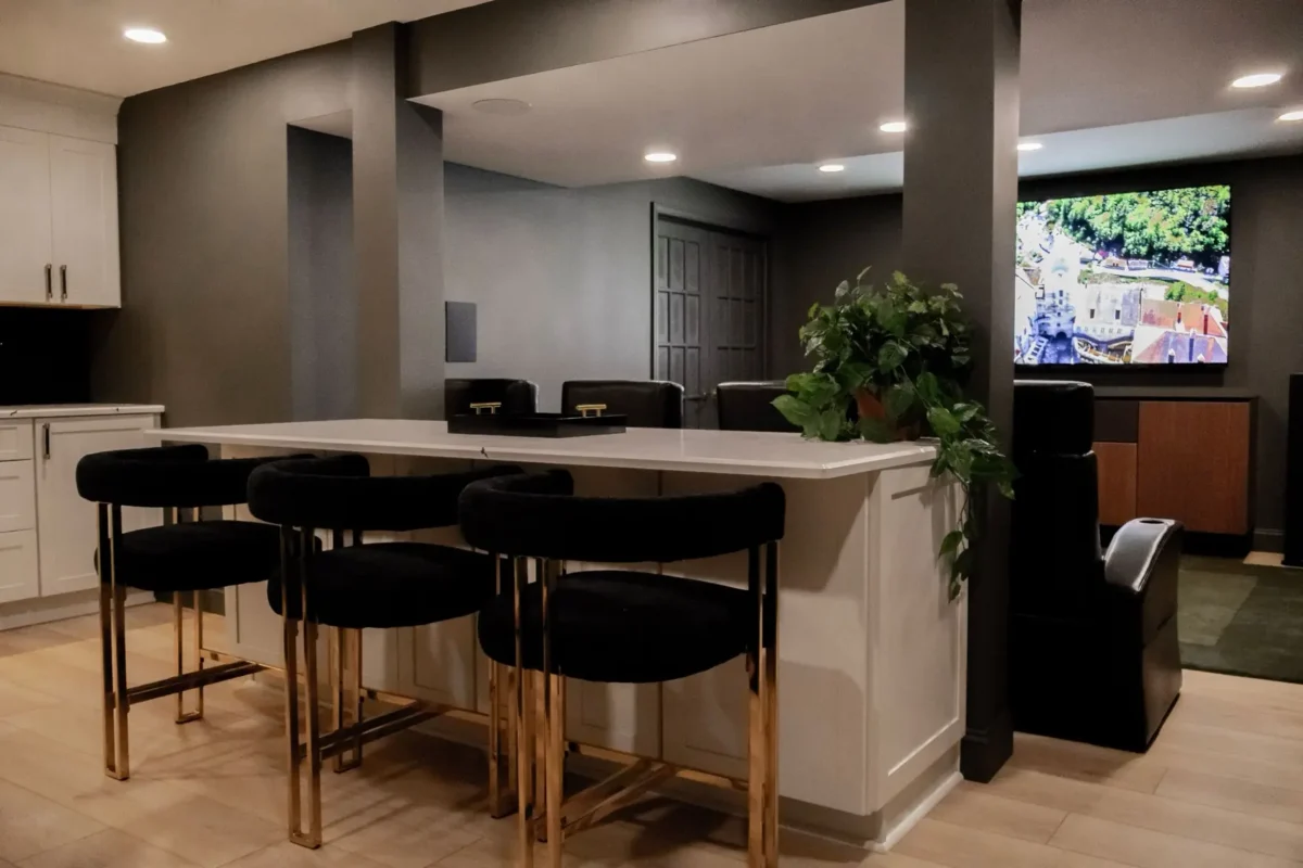 Modern basement bar with white island, black bar stools, gray walls, and a mounted TV on the right; potted plant on the counter.