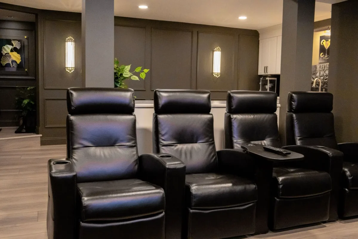 Row of four black leather recliner seats with cupholders in a dark modern home theater setting, facing away from the camera.