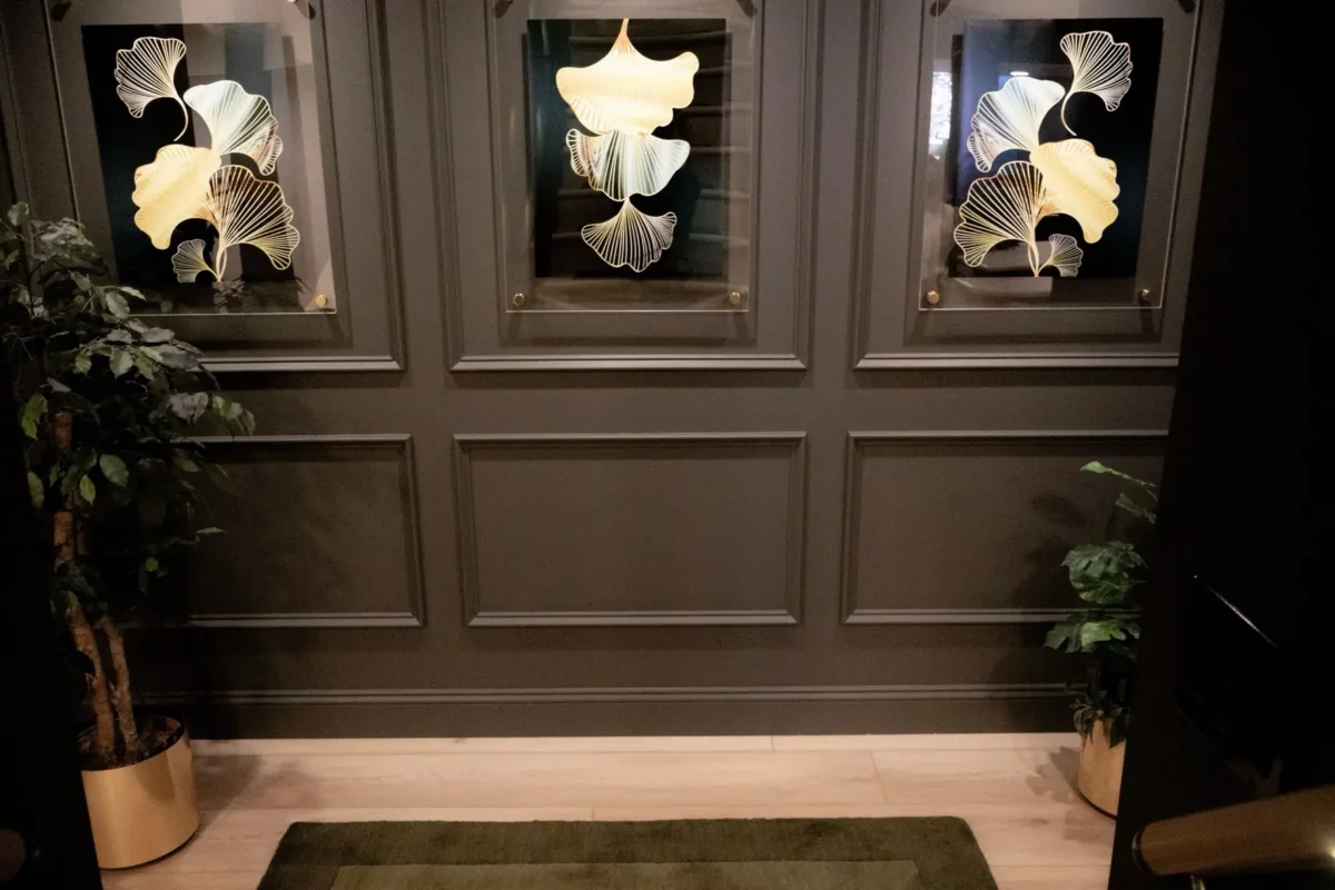 Three framed gold ginkgo-leaf artworks on a dark paneled wall, with tall potted plants in gold pots on either side in a cozy interior.