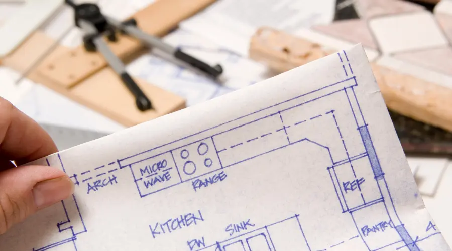 Hand holding a kitchen blueprint showing layout and labeled appliances, with drafting tools in the blurred background.