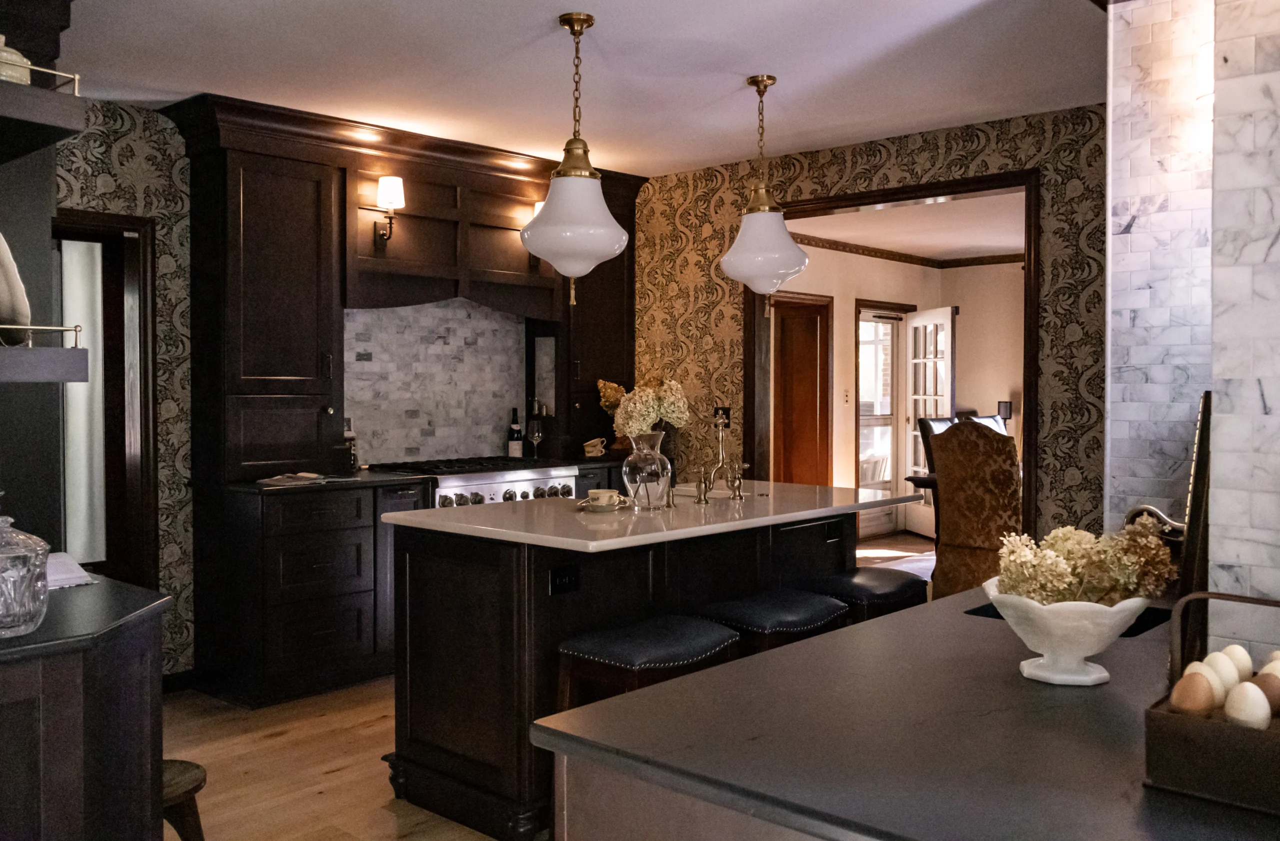 Dark wood kitchen with a white island, two glass pendant lights, floral wallpaper, and a doorway to the dining area.