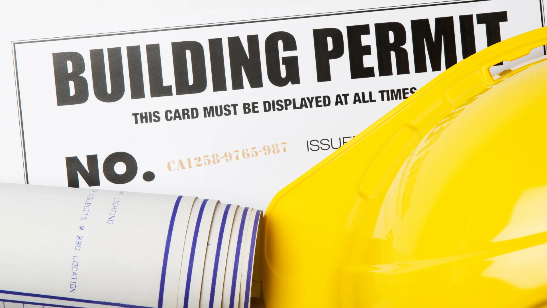 Building permit sign with a yellow hard hat and rolled blueprints on a construction site, indicating safety and compliance.