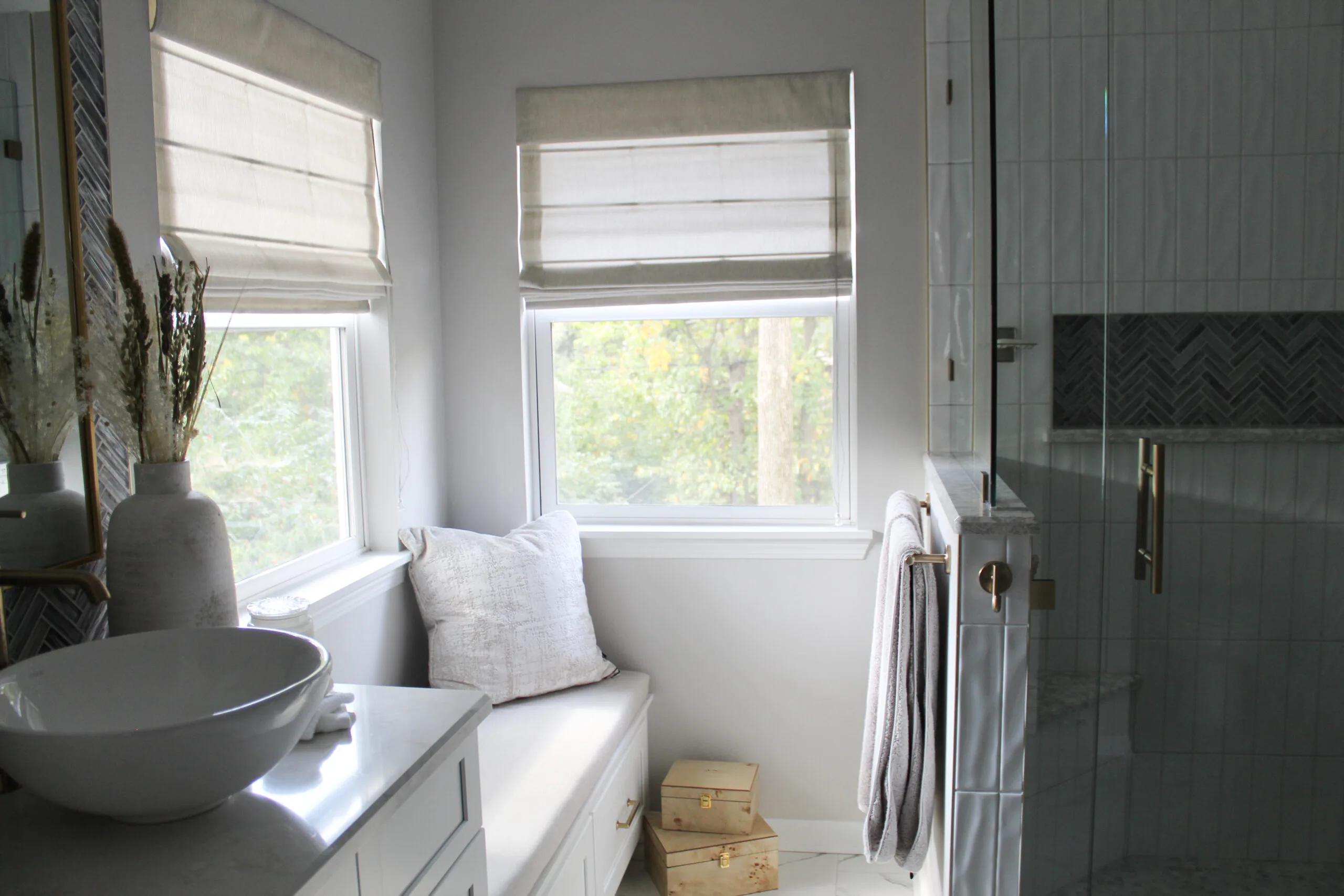 Bathroom interior with a window and beige roman shades, a white bowl sink on a marble countertop, and decorative vases nearby.
