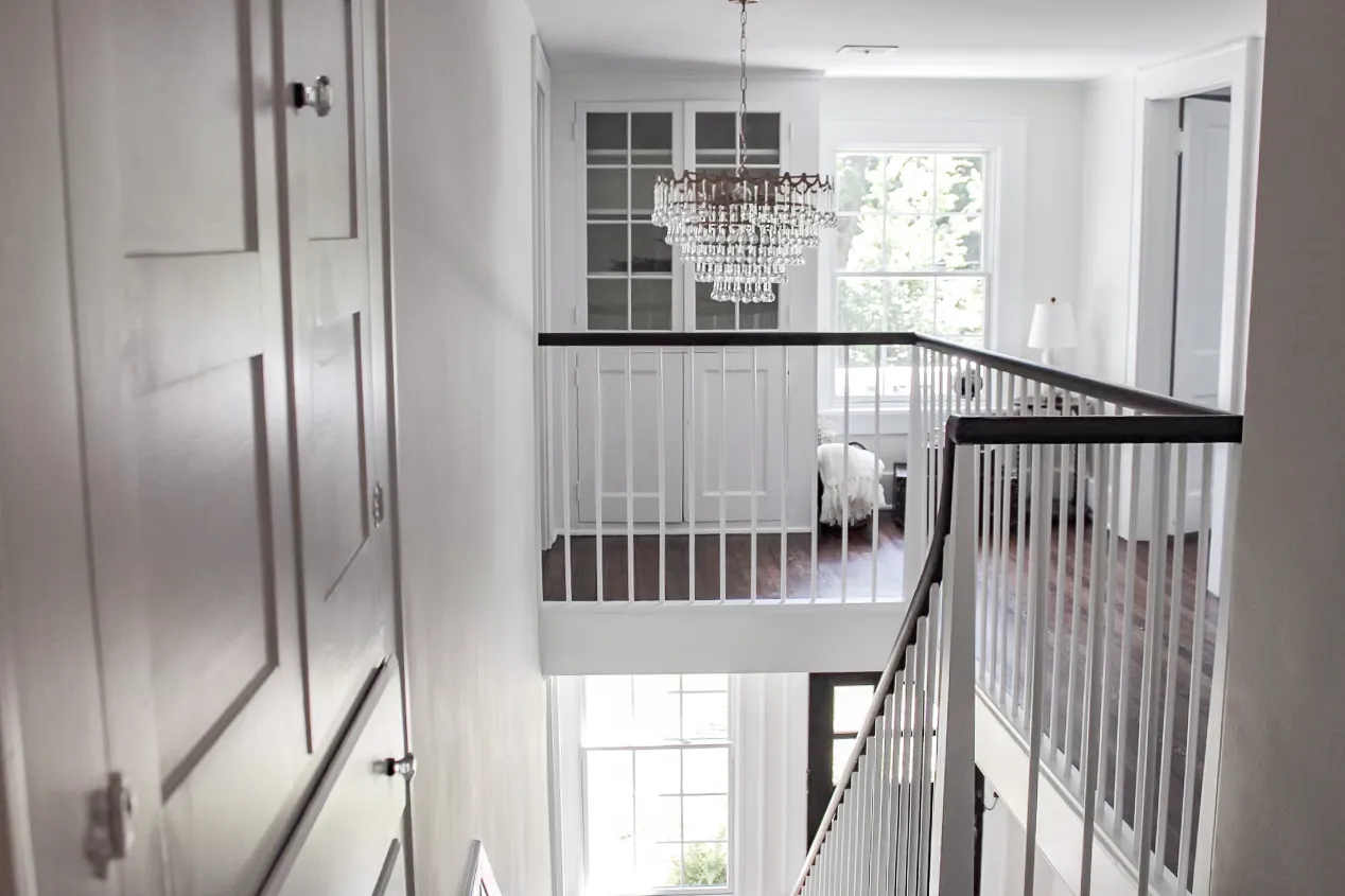 Bright two-story hallway with a crystal chandelier, white railings, and large windows letting in sunlight.
