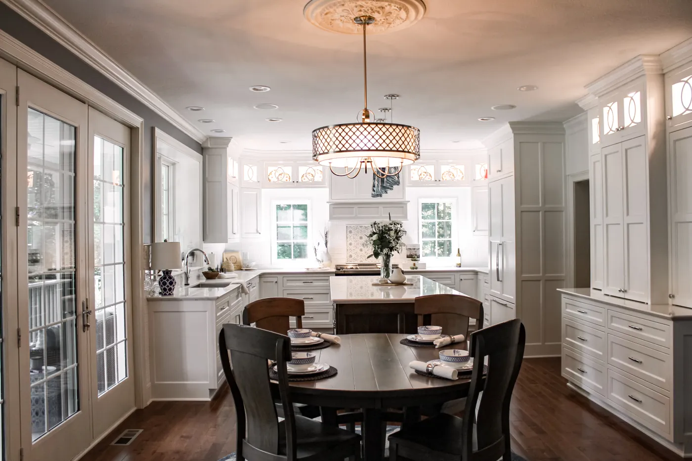 Bright white kitchen with an island, dark wood dining table for four, chandelier, and French doors on the left.