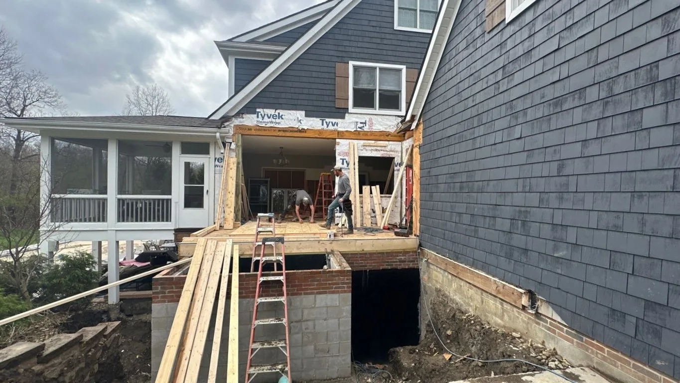 Home addition under construction with new framing and housewrap, a block and brick foundation wall below, and a finished screened-in porch beside it